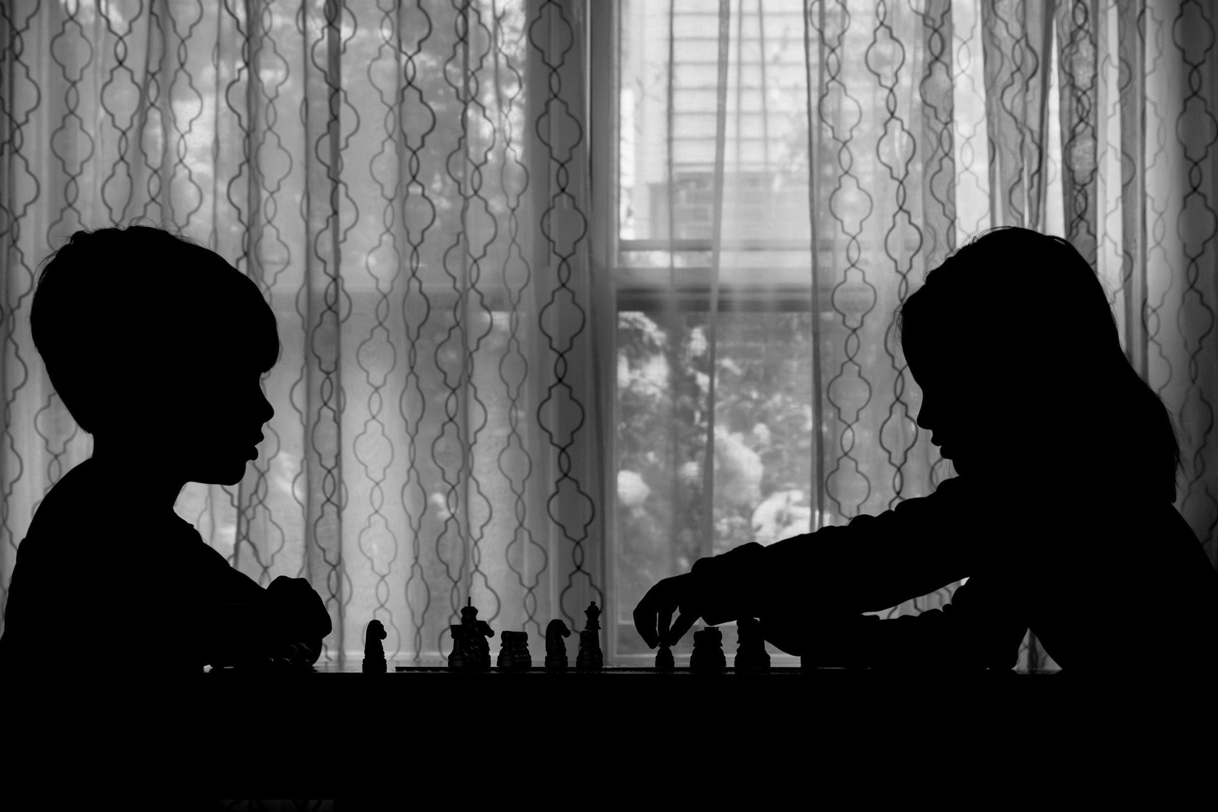Black-and-white silhouette of two children playing chess across a table as one reaches to make a move in front of a sunlit window.