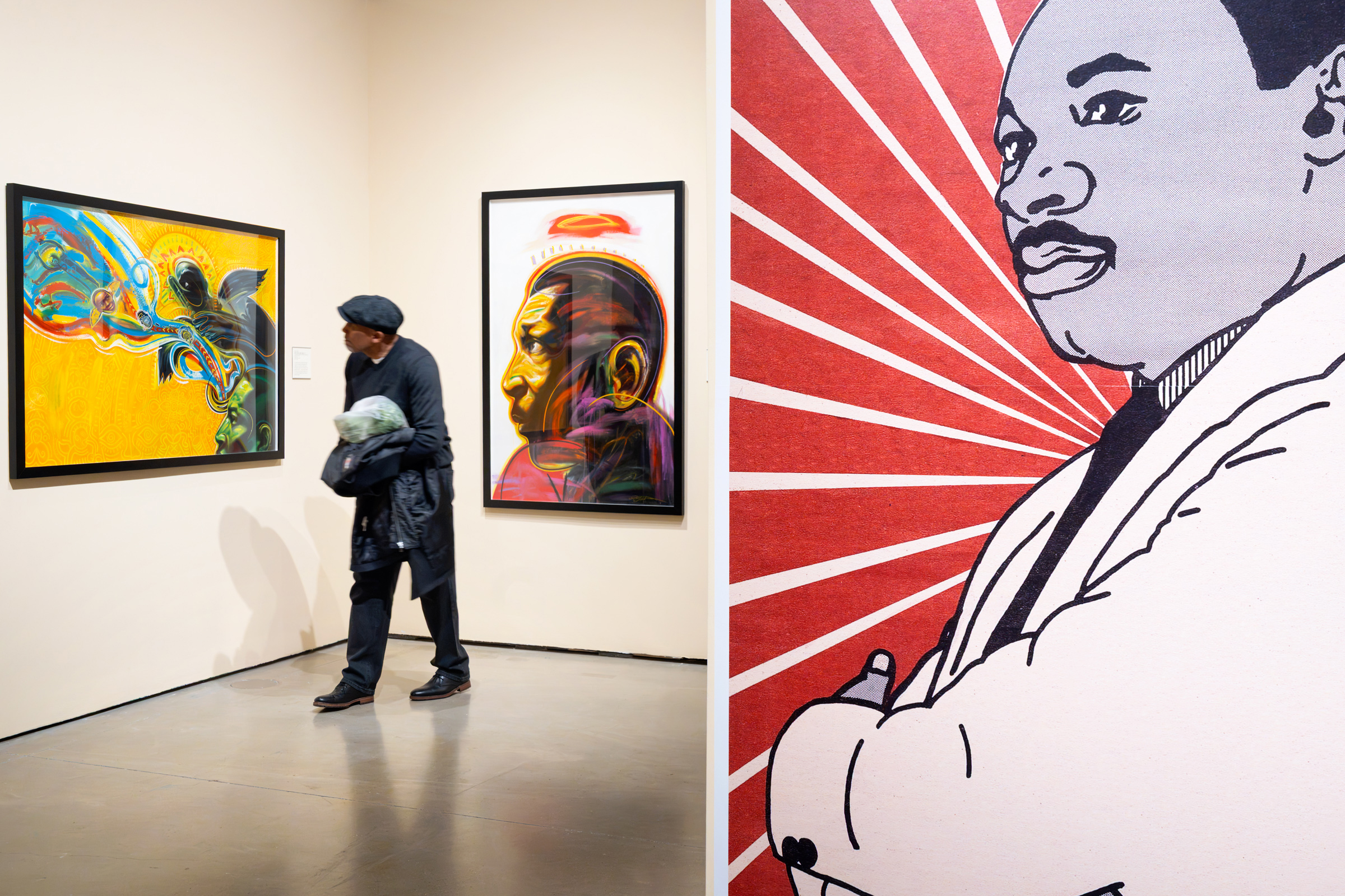 Museum visitor walks between colorful portraits during the Delaware Art Museum's Martin Luther King Jr. Day exhibition, with a large image of Dr. Martin Luther King Jr. filling the adjoining wall.