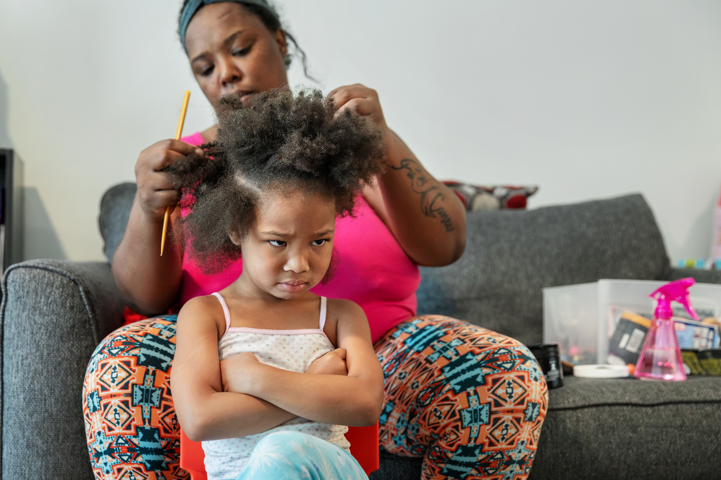 A young girl sits with her arms crossed and pouts while her mother styles her hair from the couch behind her.