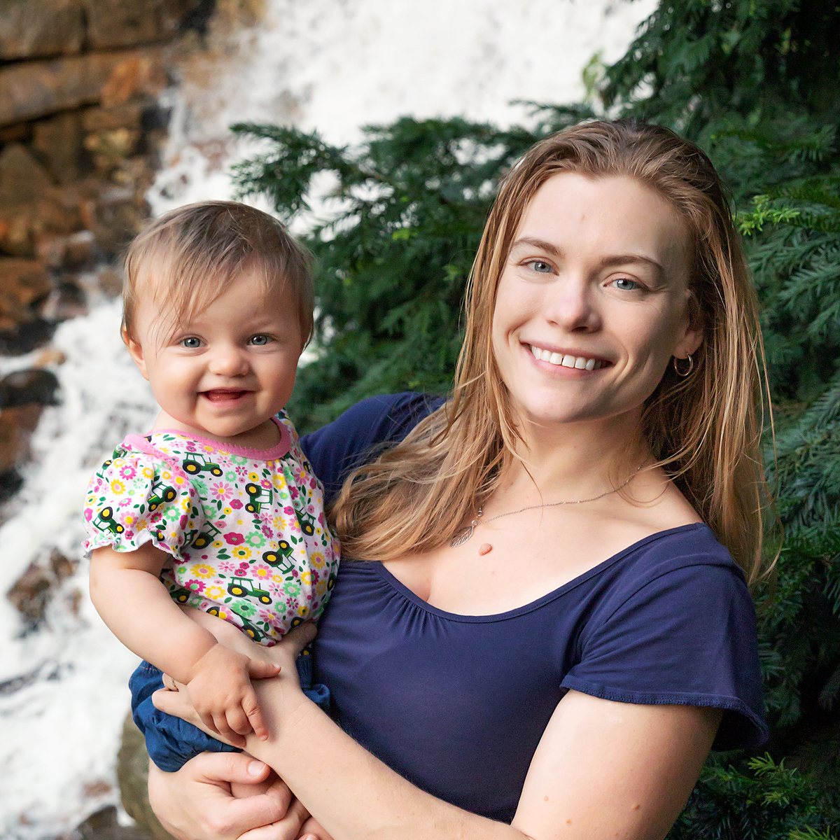 Laura and her daughter smile together as Laura holds her in front of a waterfall and evergreen trees.