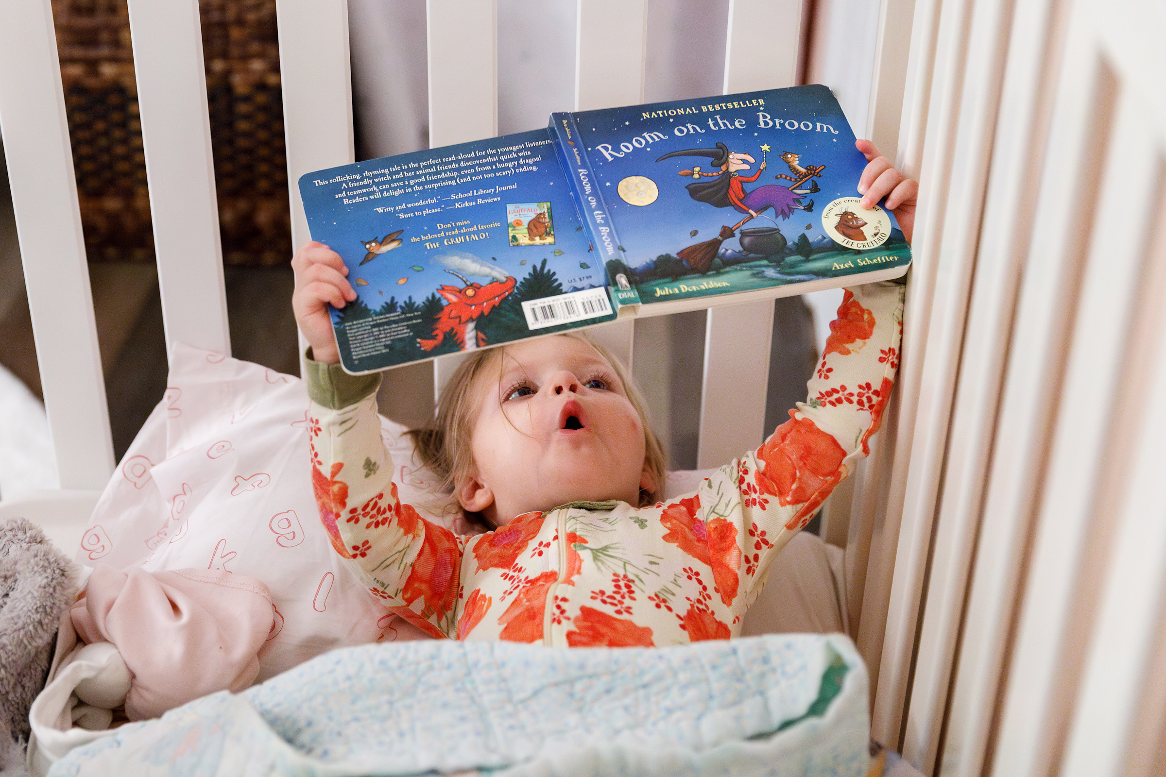 Child lying in a crib, holding an open picture book overhead while looking up in wonder during a quiet bedtime moment.