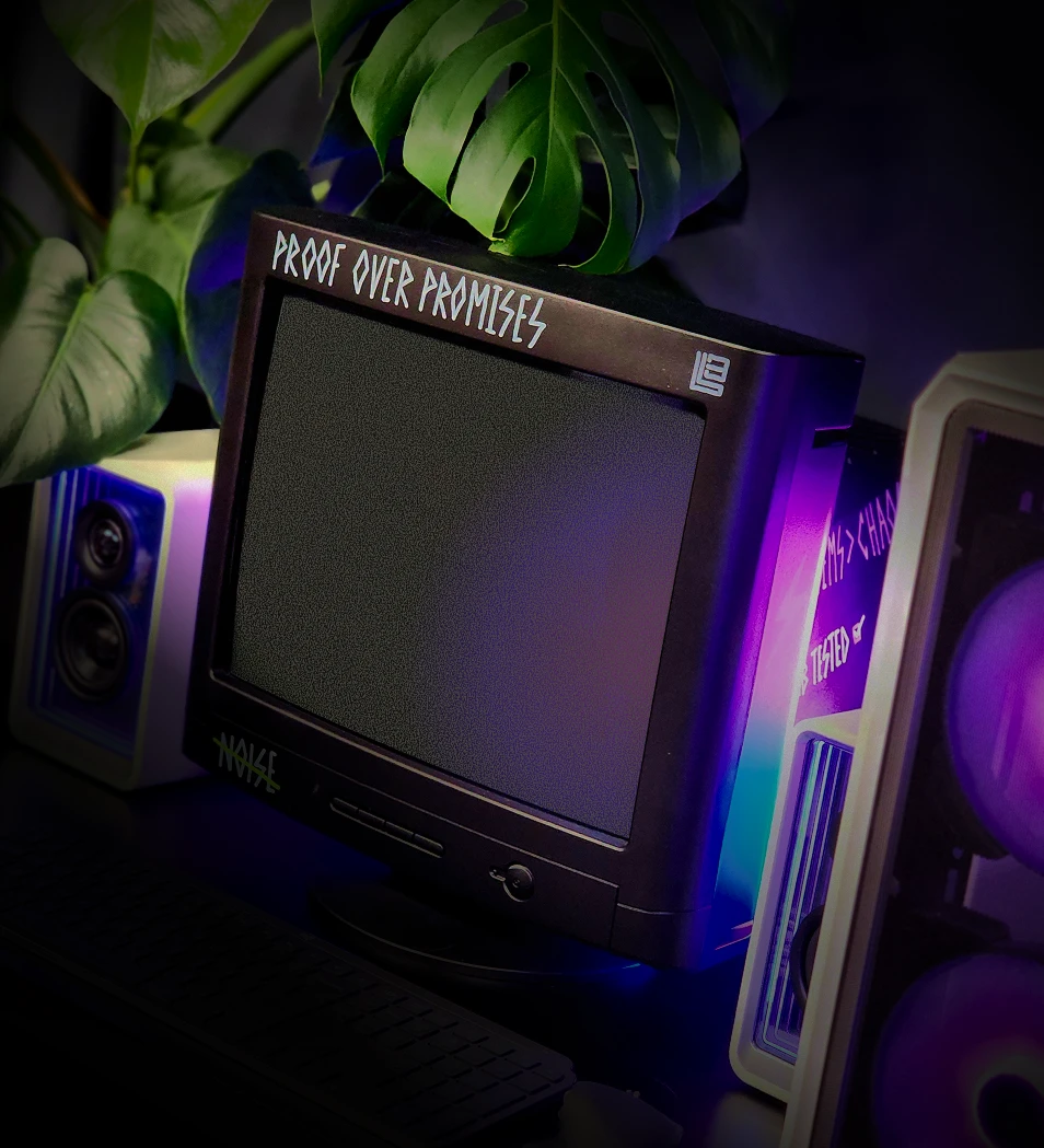 Retro black monitor with captions written on it sitting on a desk with plants and purple-lit speakers nearby.