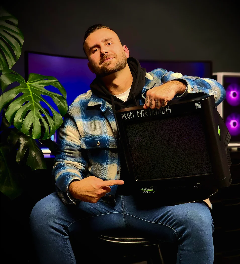 Logan in blue plaid jacket holding and pointing to an old CRT monitor in a dark room.