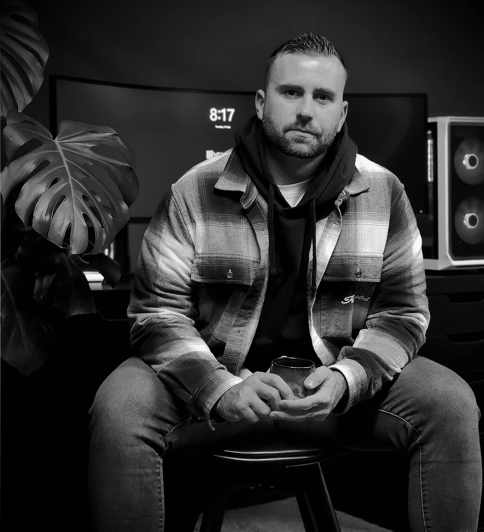 Logan in plaid shirt and hoodie sitting on stool holding a cup with computer screens and plant behind.