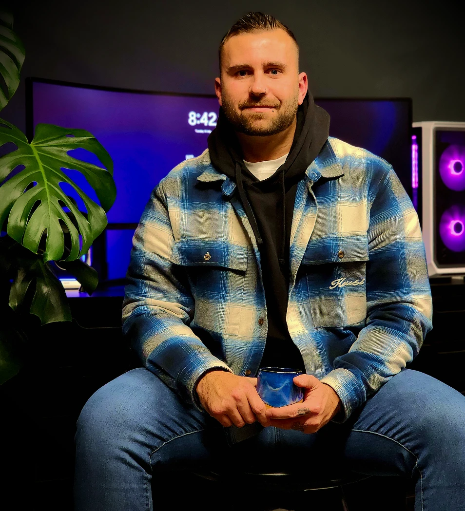 Man in blue plaid jacket and jeans sitting with a cup in hand in front of a curved monitor and purple-lit device.