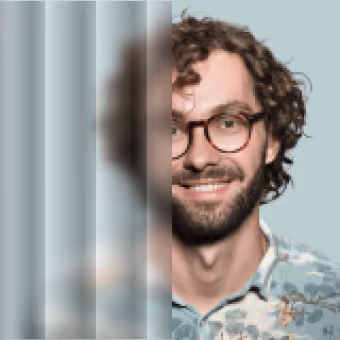Portrait of a smiling man with curly hair, beard, glasses, and a patterned shirt on a light blue background.