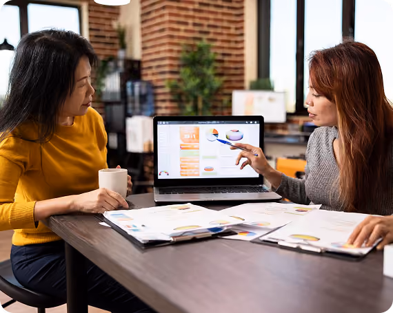 Two women at a table discussing charts on a laptop and printouts in a modern office.