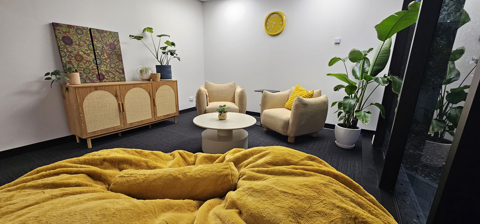 Cozy sitting area with beige armchairs, yellow cushions, round table, indoor plants, and wooden cabinet with art.