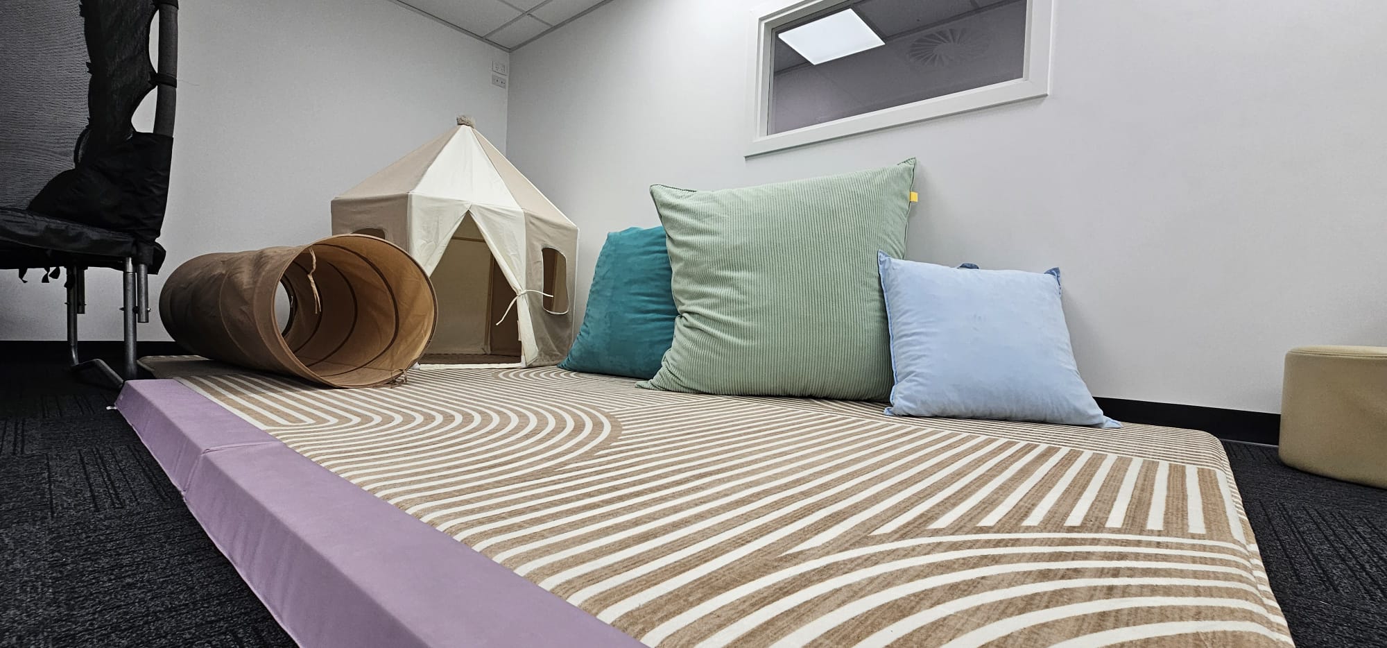 A play area with a patterned mat, three cushions, a fabric tunnel, and a small tent in a room.