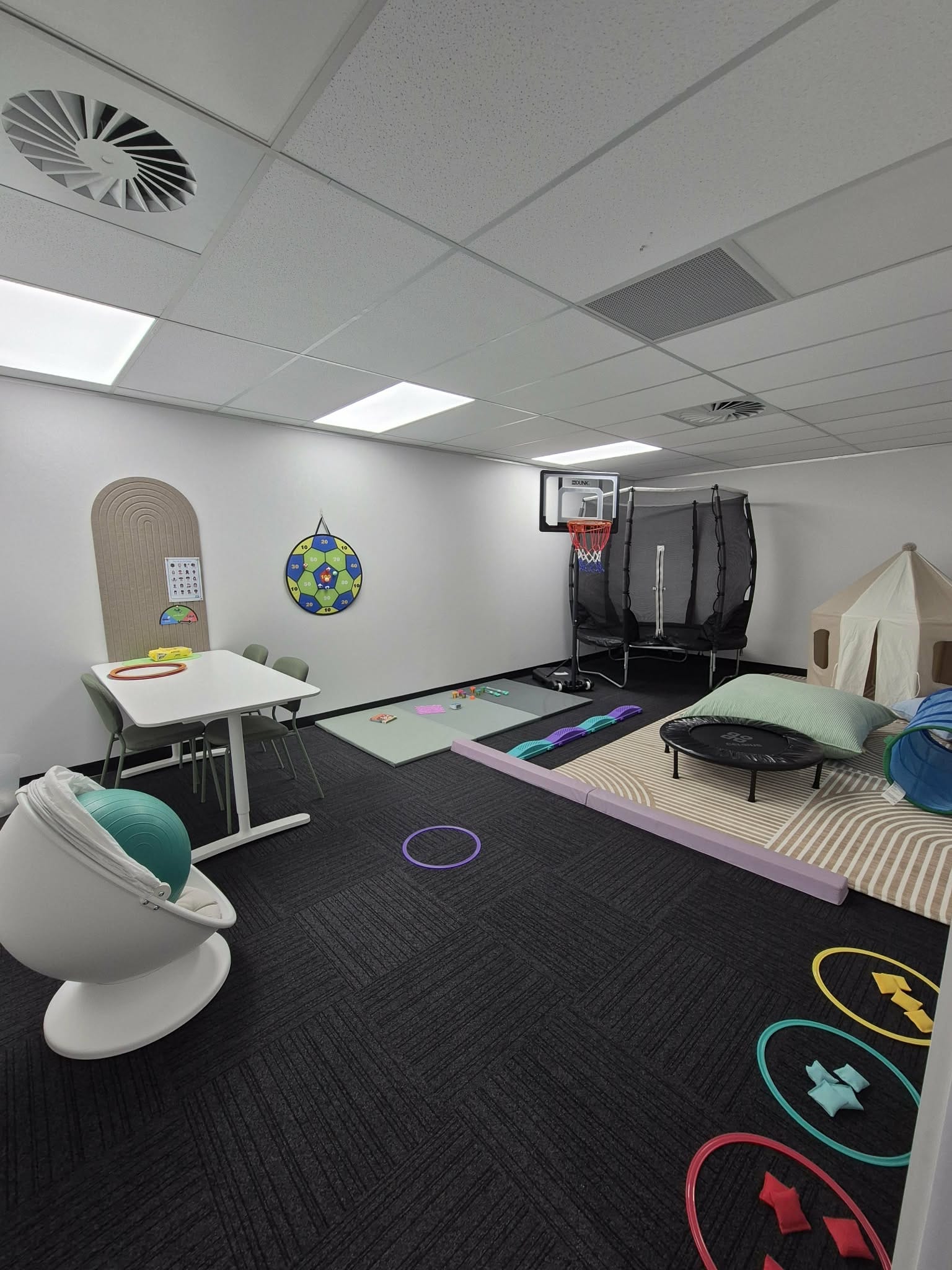 Playroom with tables, chairs, trampoline, small basketball hoop, play tent, cushions, and colorful rings on dark floor.