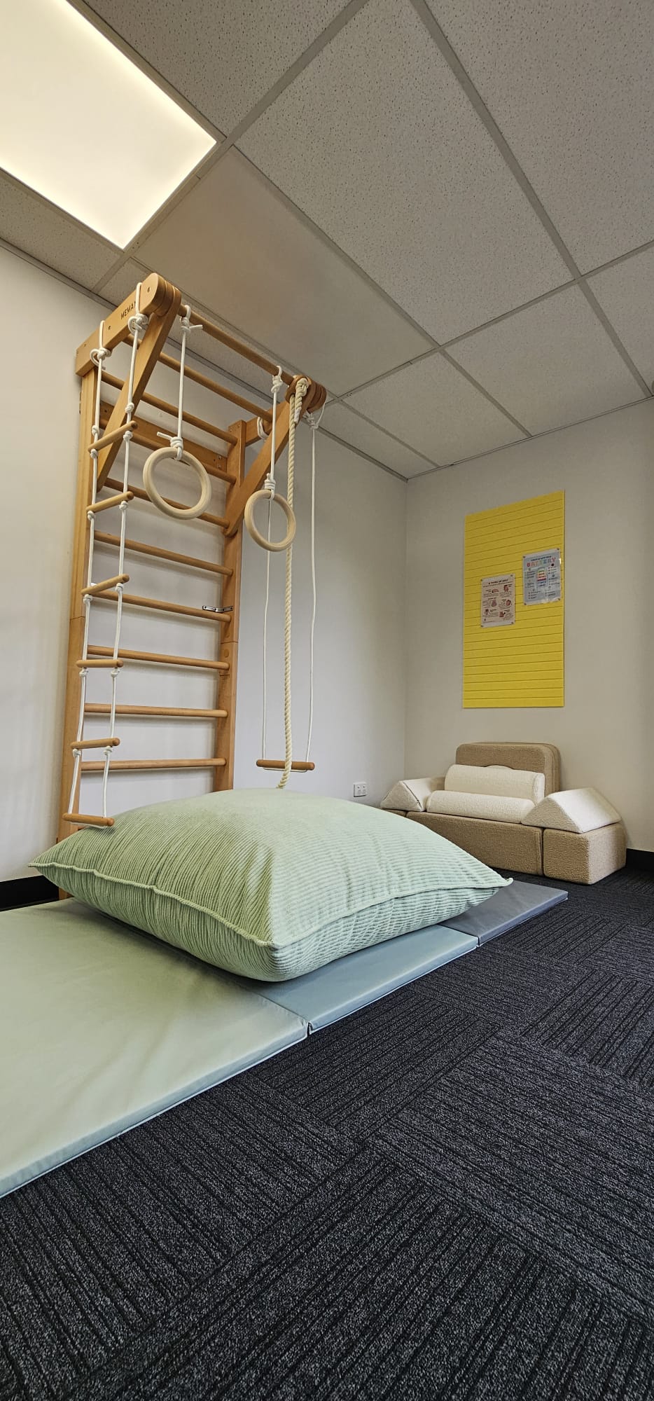 Indoor play area with wooden climbing structure, gym rings, rope swing, cushion, and soft seating.