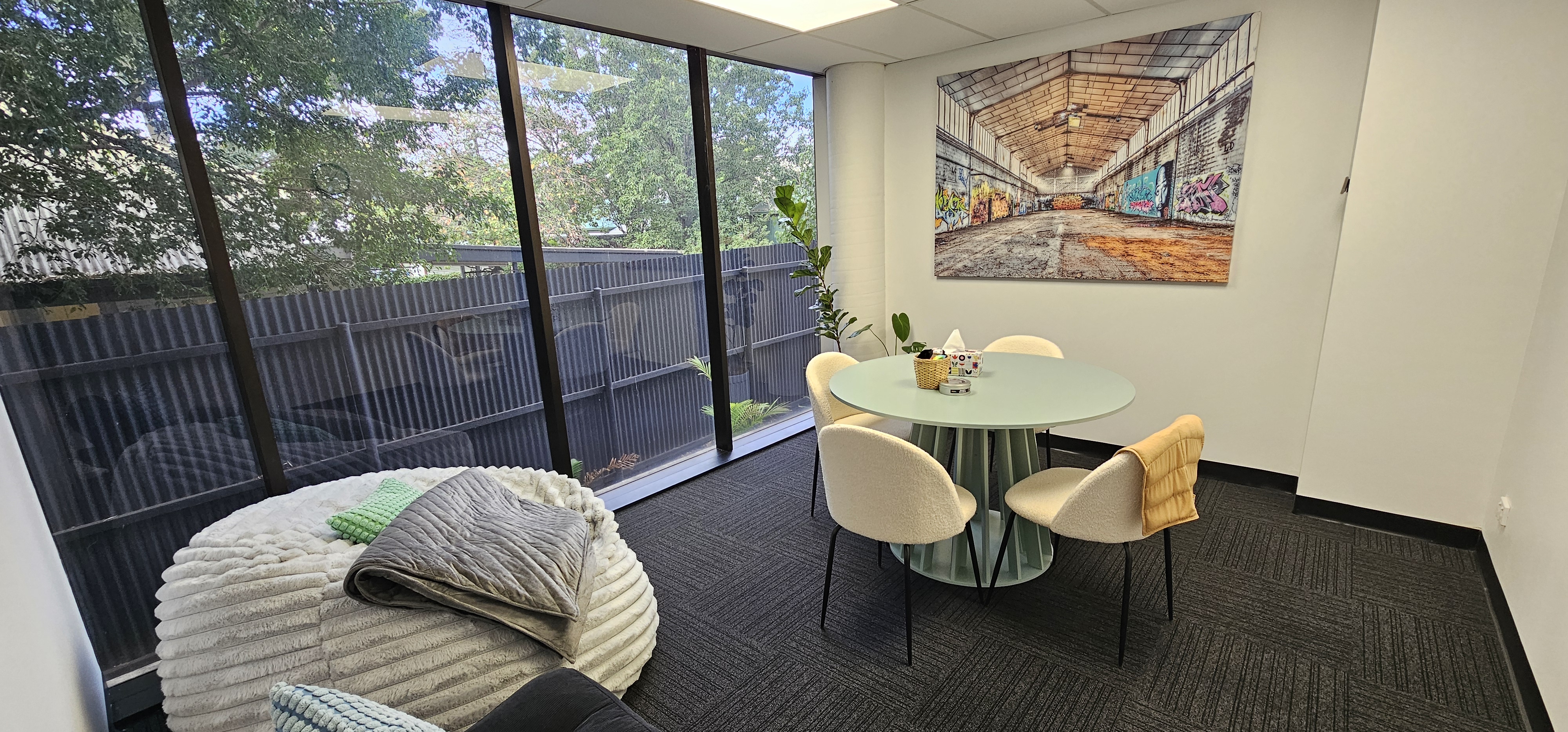 Small meeting room with round table, four chairs, large window, cushions, and plant under graffiti art.