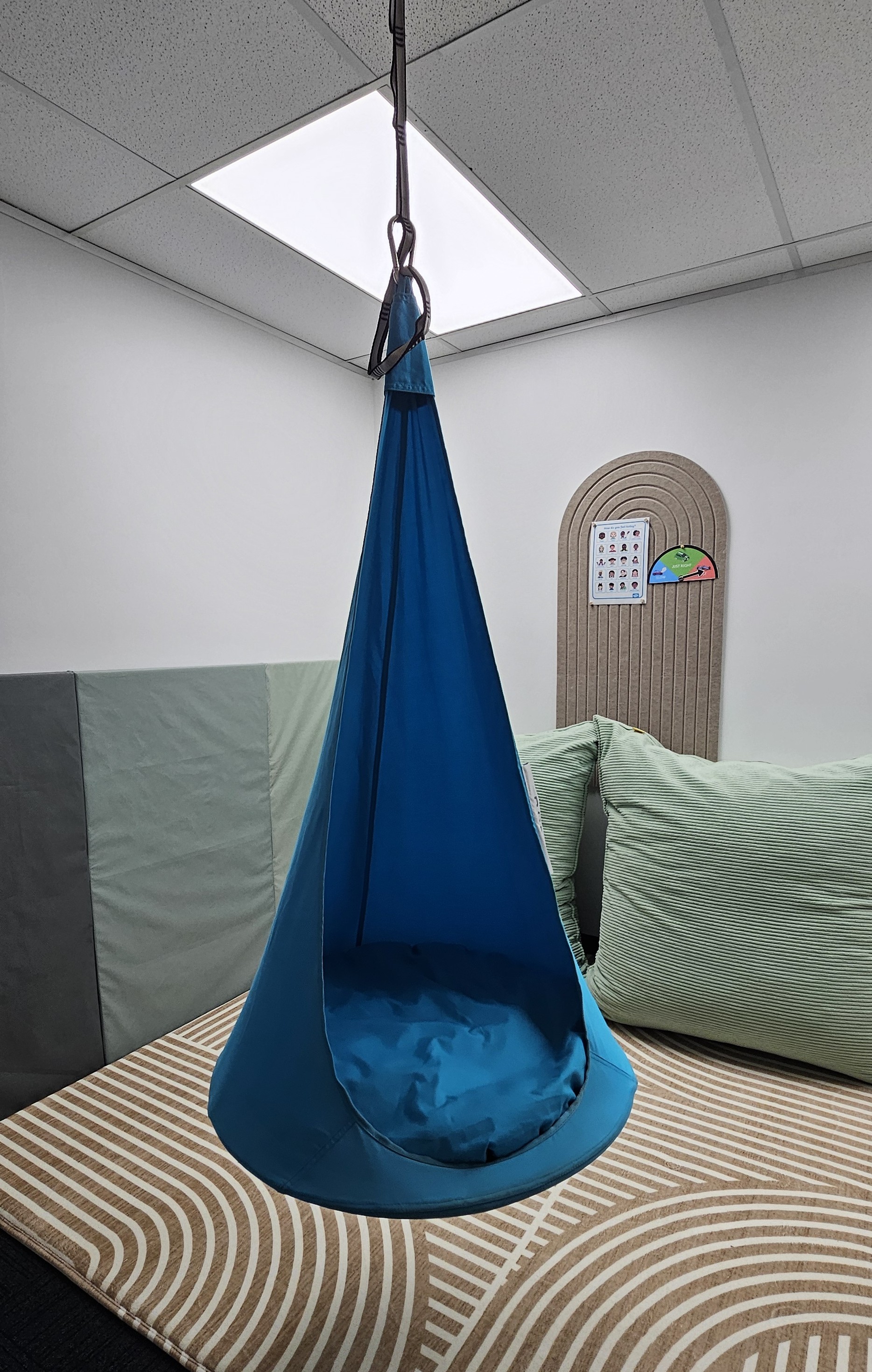 Blue hanging fabric pod chair suspended indoors over patterned beige rug with cushions nearby.