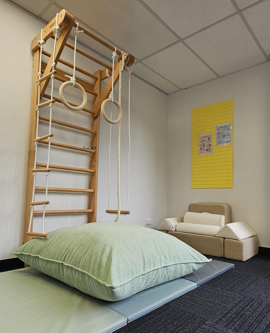 Indoor play area with wooden climbing structure, gym rings, rope swing, cushion, and soft seating.