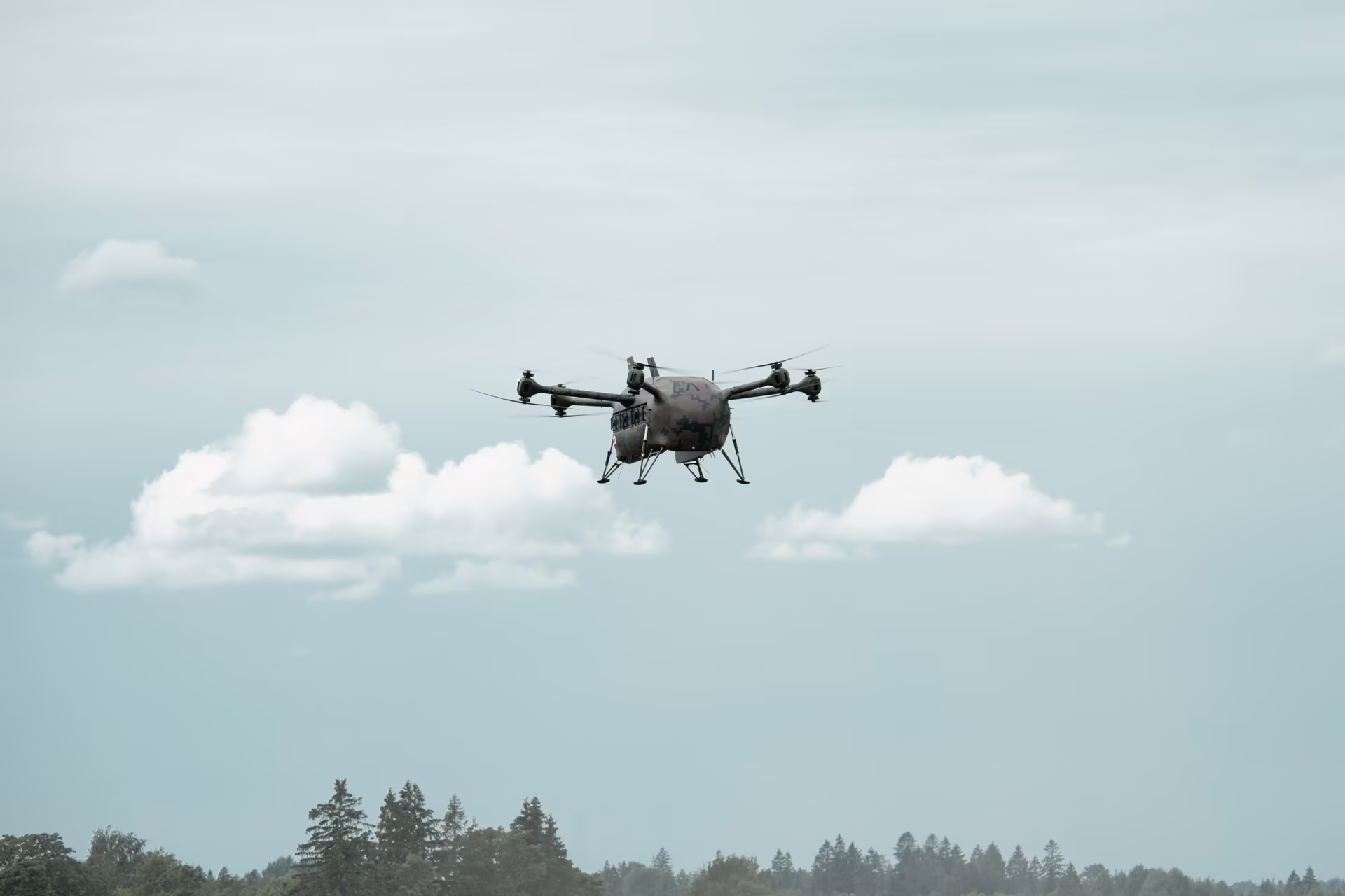 Grille drone flying above a treeline under a cloudy sky