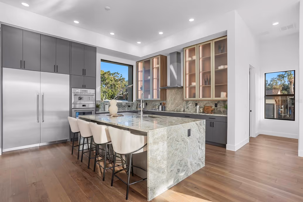 Modern kitchen with a large marble island, four upholstered bar stools, grey cabinets, stainless steel refrigerator, built-in oven, and hardwood floors.