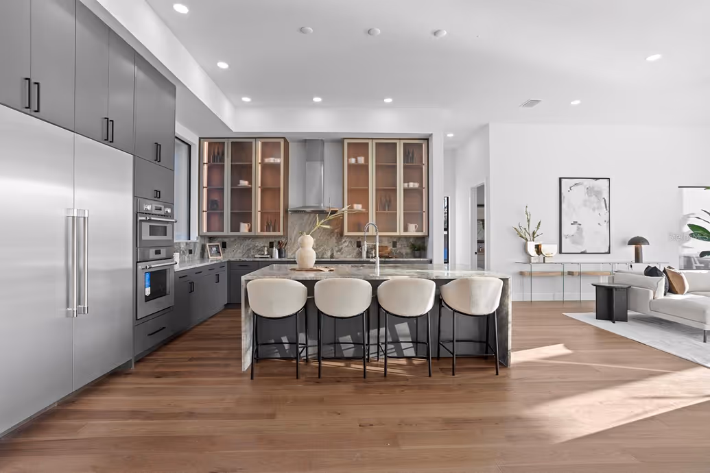 Modern kitchen with dark gray cabinets, stainless steel appliances, a marble island with four white cushioned chairs, and wood flooring.