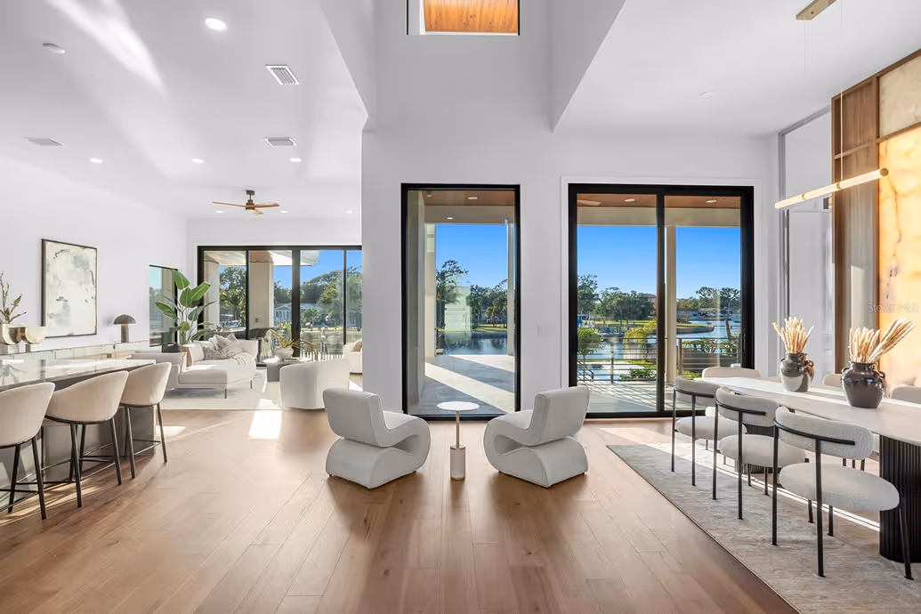 Modern open living space with light wood floors, two sculptural white chairs facing floor-to-ceiling windows overlooking a lake, and adjacent dining and lounge areas.