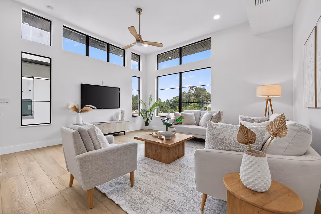 Bright modern living room with large windows, beige sofas, wooden coffee table, and ceiling fan.