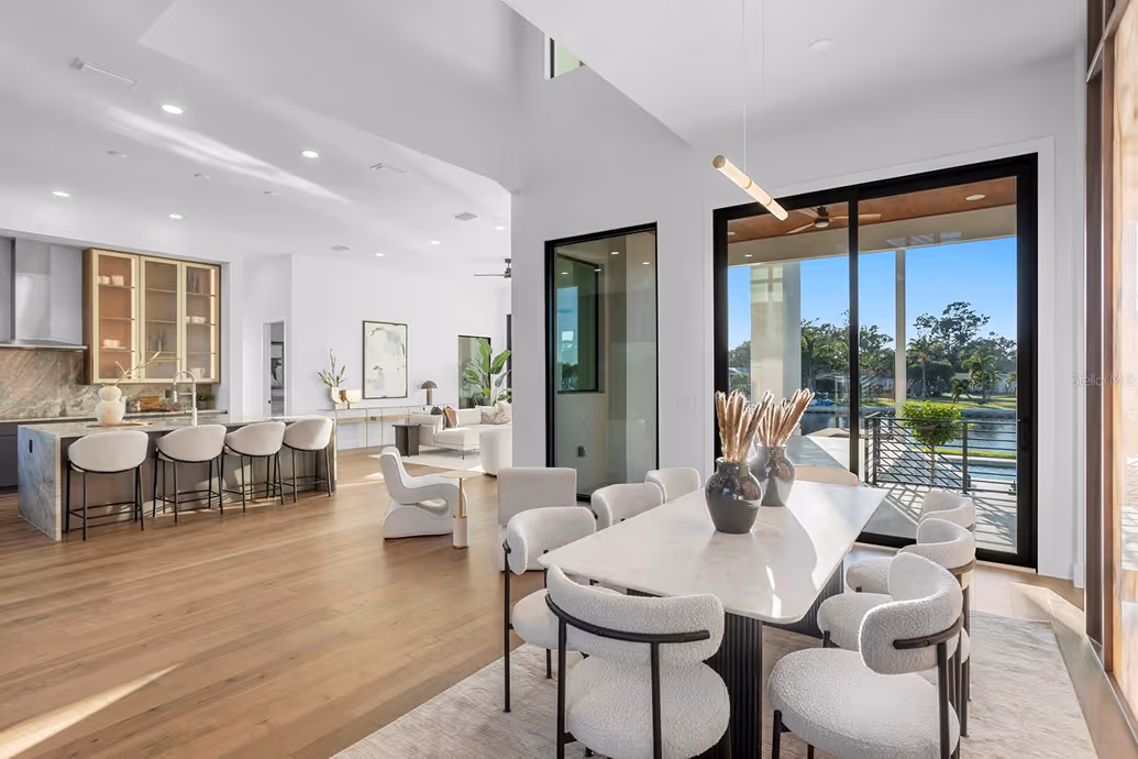 Modern open dining and kitchen area with white cushioned chairs, marble dining table, large sliding glass doors overlooking a patio and water view.