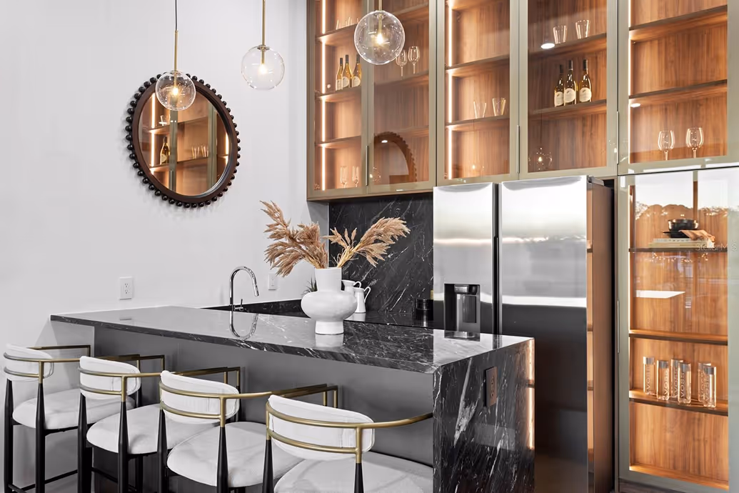 Modern kitchen with black marble island, four white cushioned bar stools, hanging glass pendant lights, round mirror, and wooden cabinets with glass doors displaying glassware and bottles.