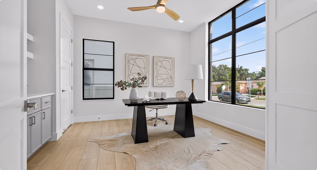 Modern office with black desk, rolling chair, lamp, wall art, large window, and ceiling fan.