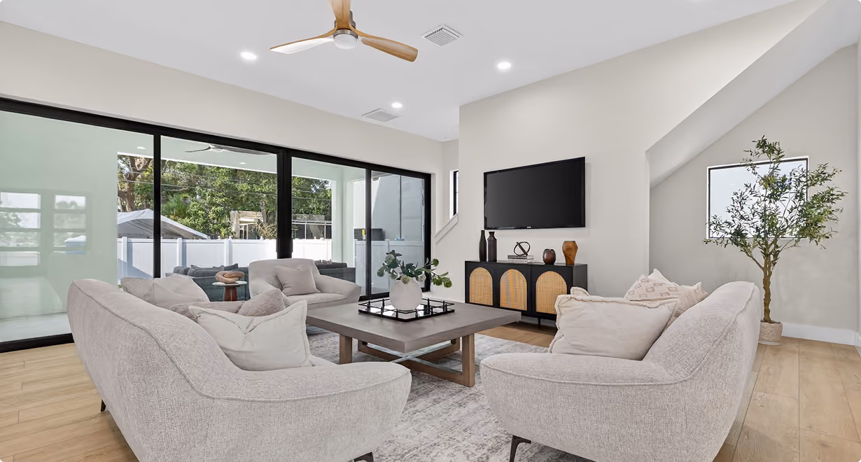 Modern living room with beige sofas, coffee table, TV mounted on wall, large glass doors, and a potted plant.