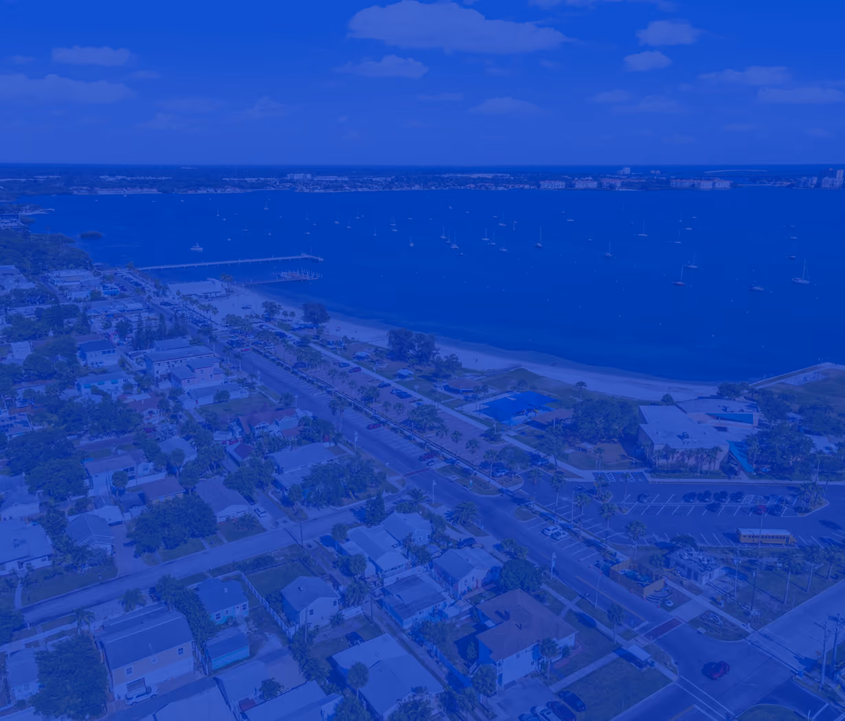 Aerial view of a coastal town with houses, a beach, marina, and boats on calm water under a blue sky.