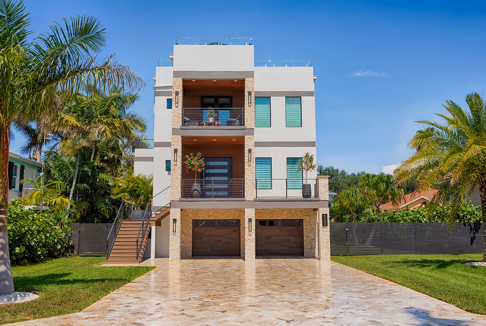 Modern three-story house with balconies, palm trees, and long tiled driveway under a clear blue sky.