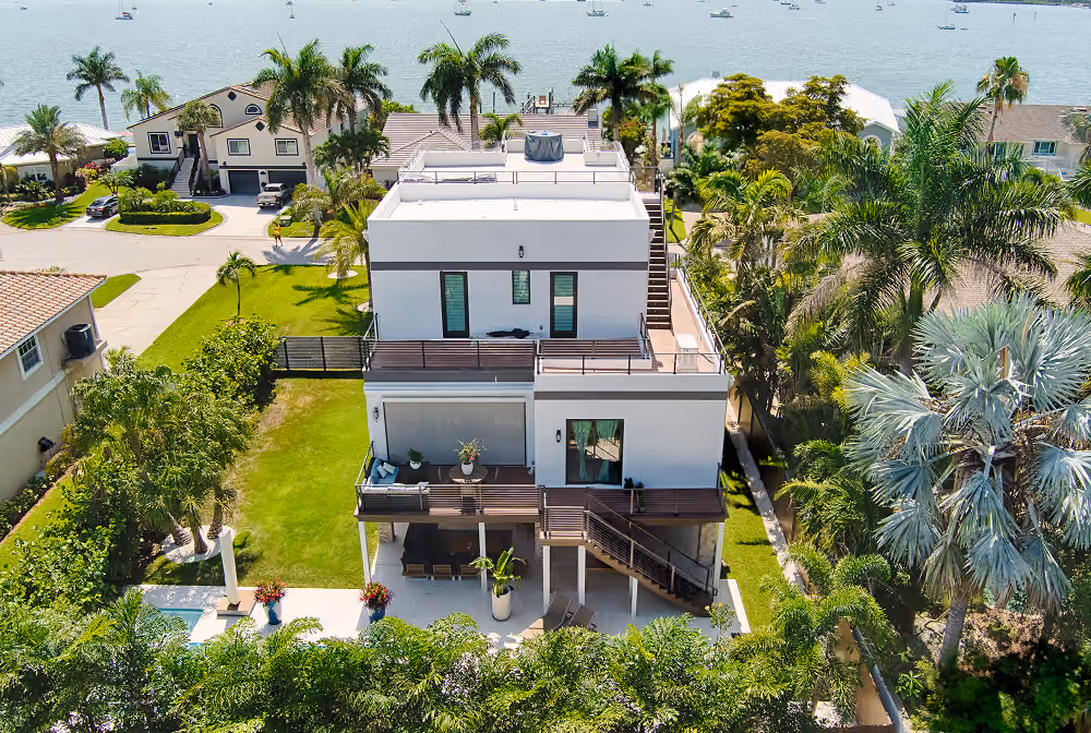 Modern white multi-story waterfront house with balconies and palm trees by a body of water with boats.