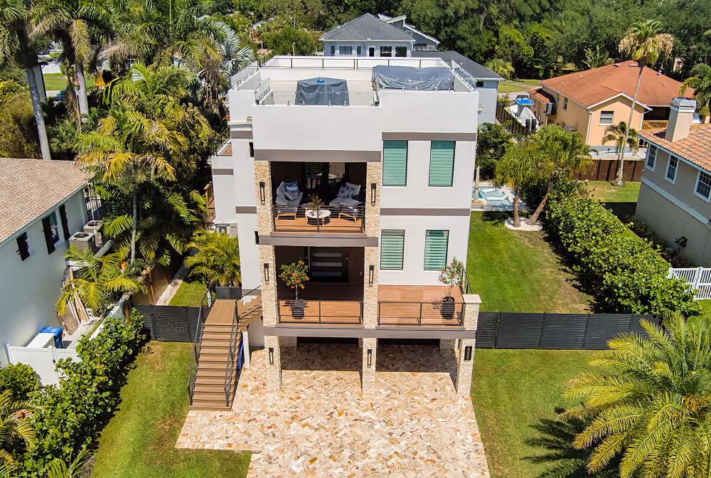 Modern three-story house with balconies, glass railing, and surrounded by green lawns and neighboring homes.