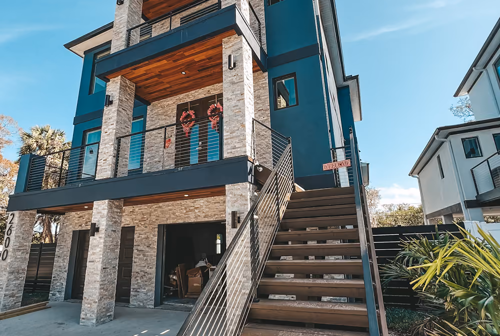 Modern two-story house with stone pillars, wood ceiling, balcony, and exterior staircase leading to second floor.
