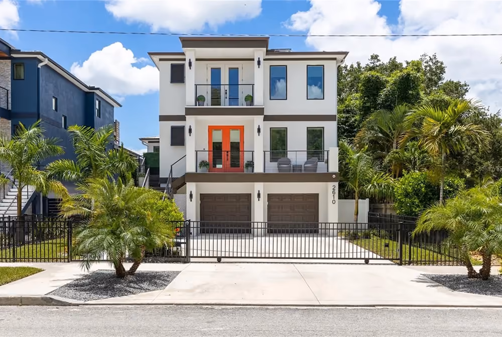 Modern three-story white house with double garage, orange door, balconies, palm trees, and metal gate.