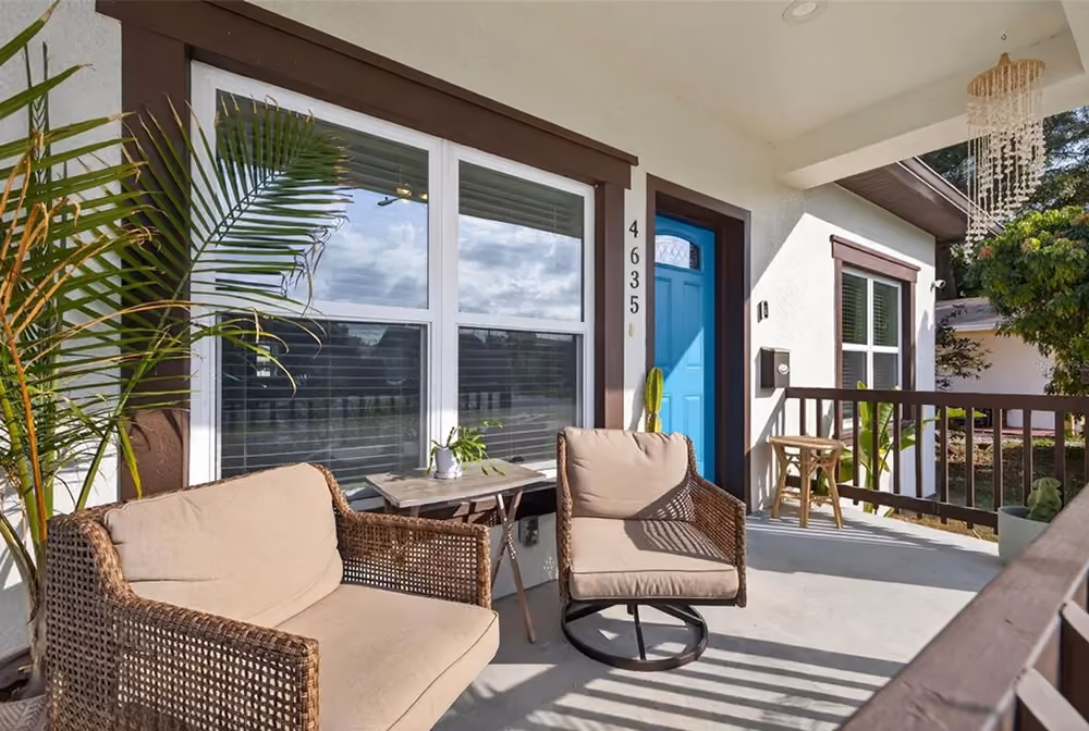 Front porch with brown wicker chairs, beige cushions, small table, blue door, and potted plants.