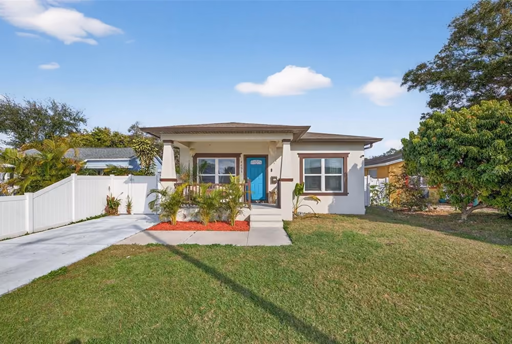 Single-story house with a blue front door, front porch, green lawn, and white fence under blue sky.