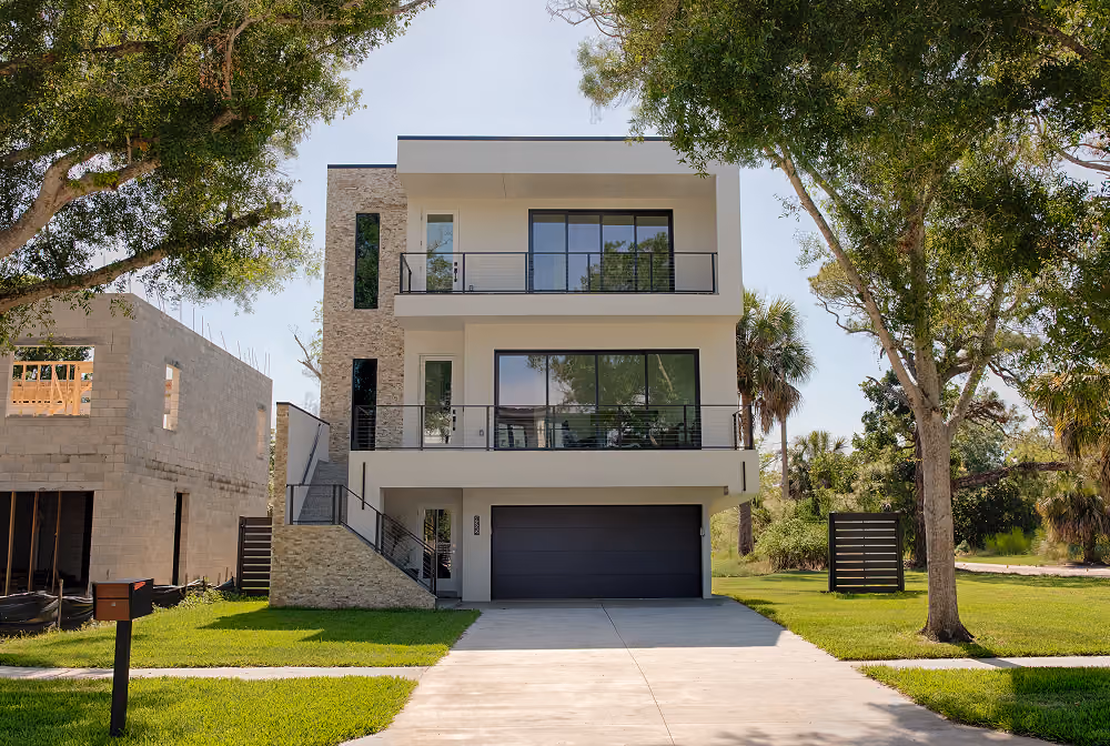 Modern three-story house with large windows, stone accents, and a driveway surrounded by trees and grass.