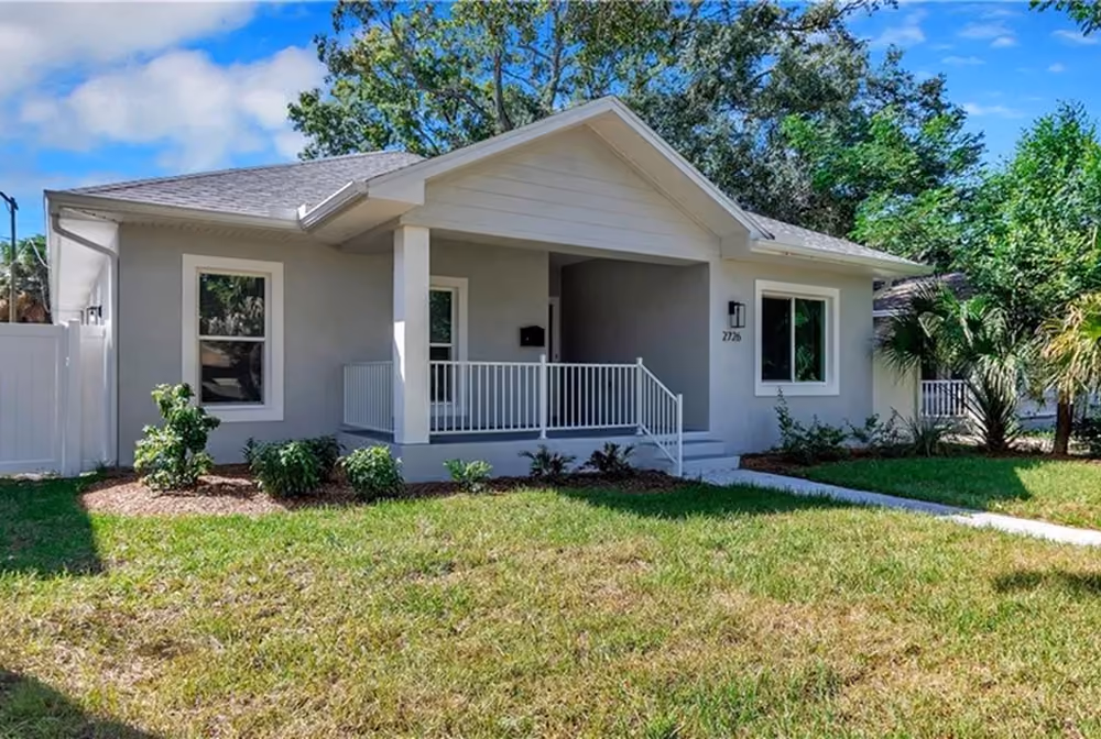 Single-story gray house with a front porch, white railing, green lawn, and trees in the background.