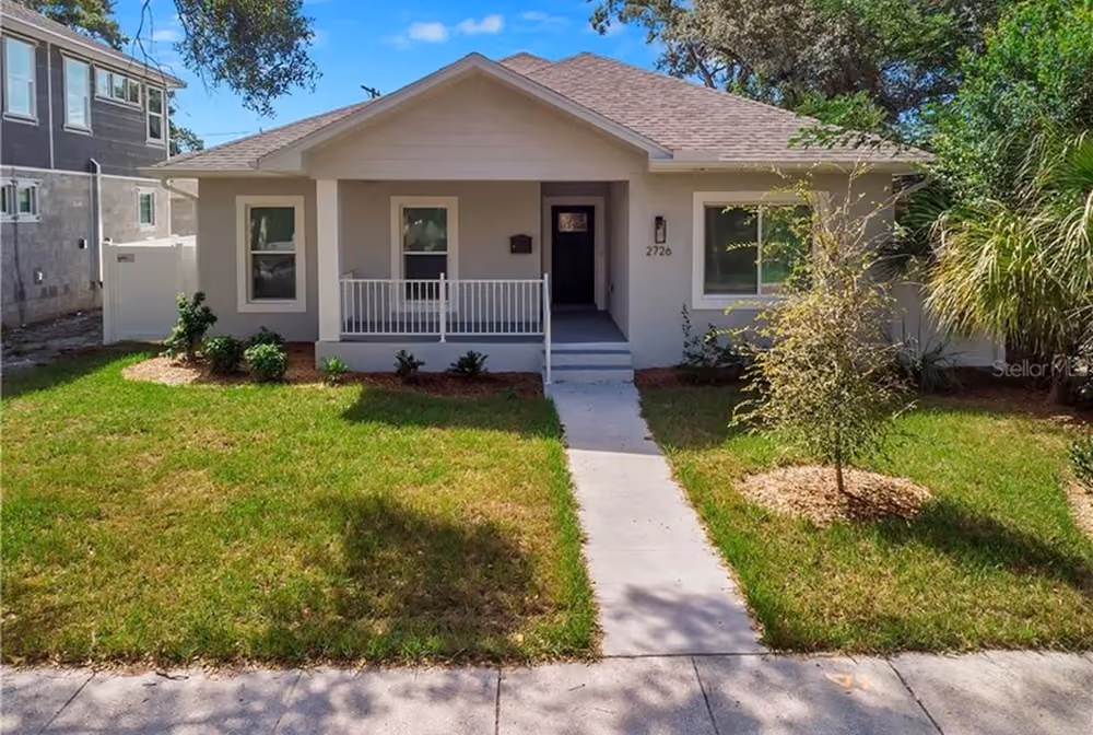 Single-story suburban house with front porch, lawn, walkway, and small trees under a blue sky.