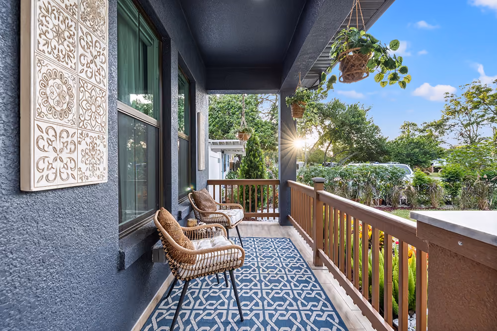 Front porch with wicker chairs, patterned rug, hanging plants, and sun setting behind trees.