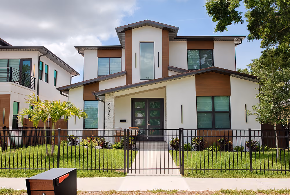 Modern two-story house with large windows, brown and white facade, and black steel fence around front yard.