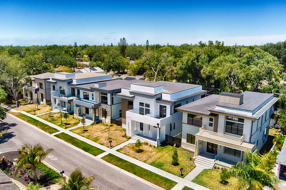A row of modern two-story homes with flat roofs, front lawns, and a tree-lined street under a blue sky.