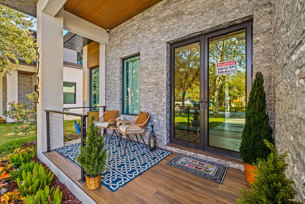Front porch with glass double doors, two wicker chairs on a patterned rug, and potted plants.