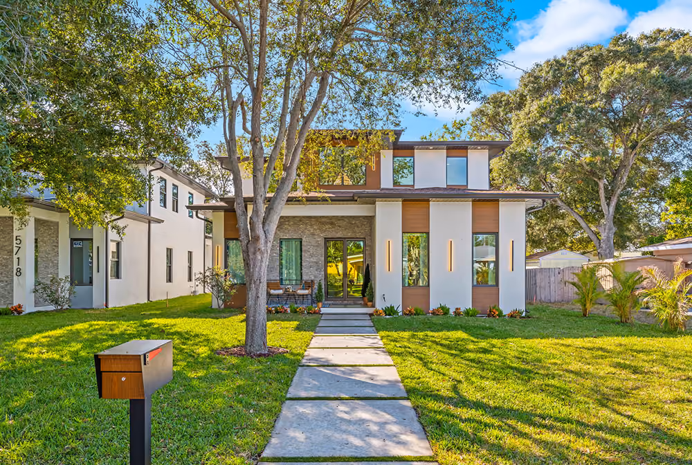 Modern two-story house with a paved walkway, large trees, and a mailbox on a sunny day.