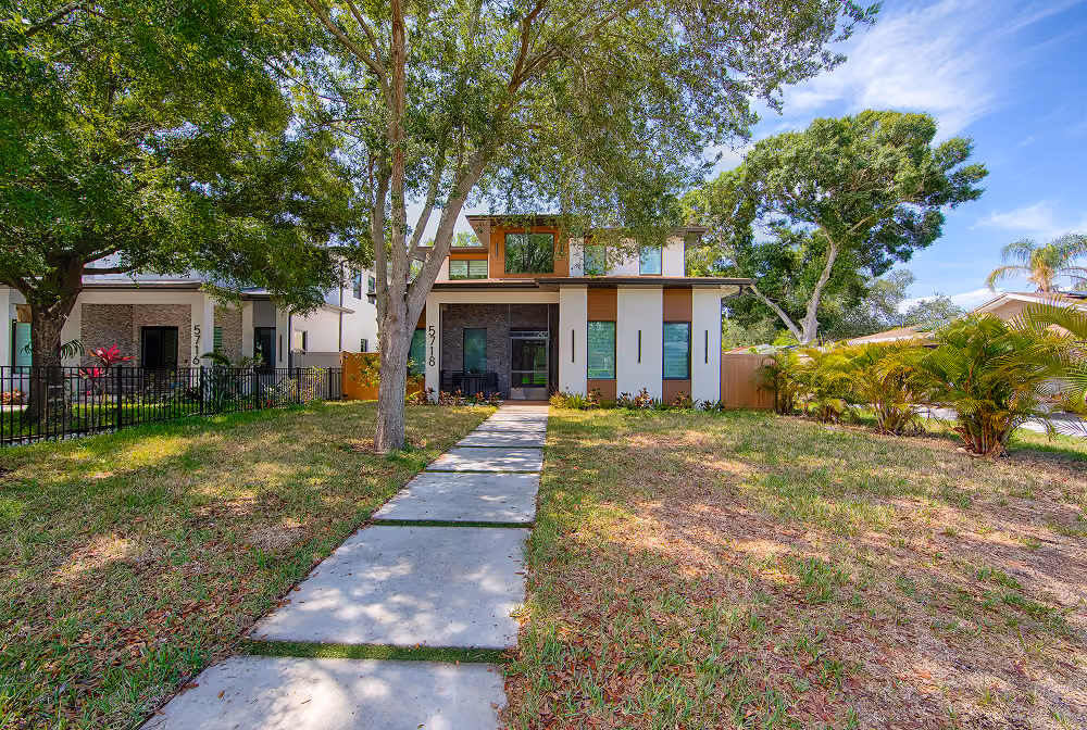 Modern two-story house with large windows, concrete path, and trees in the front yard.
