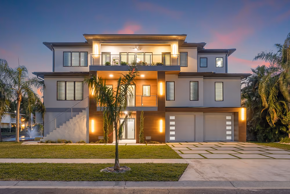 Modern three-story house with illuminated balcony, large garage, palm trees, and landscaped lawn at dusk.