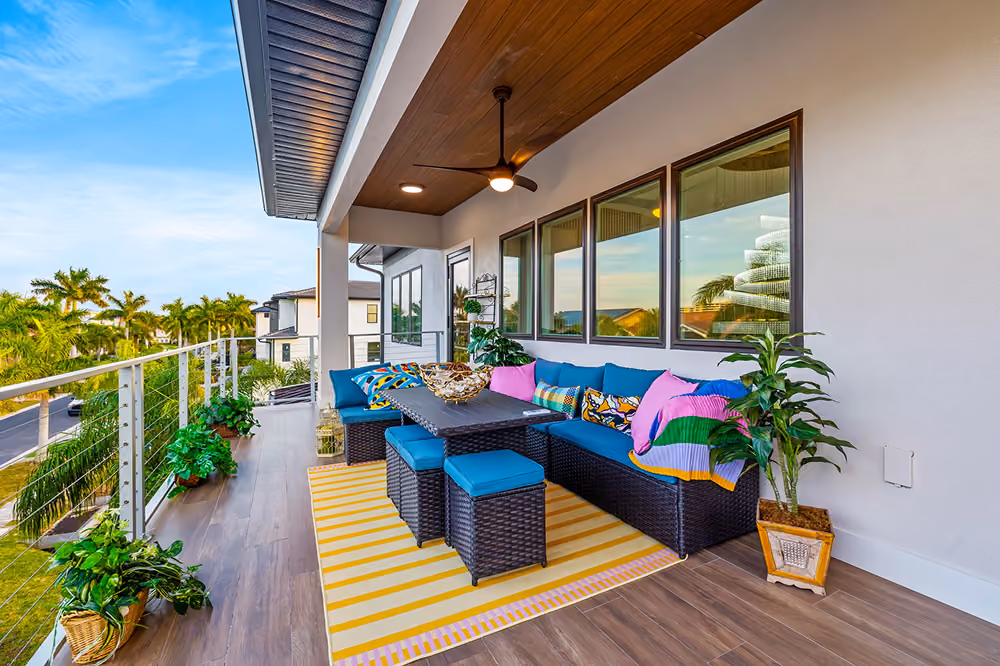 Covered balcony with blue cushioned wicker sofa, colorful pillows, yellow striped rug, and potted plants.