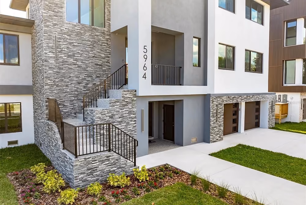 Modern multi-story house with stone accents, exterior stairs, garage doors, and landscaped front yard.