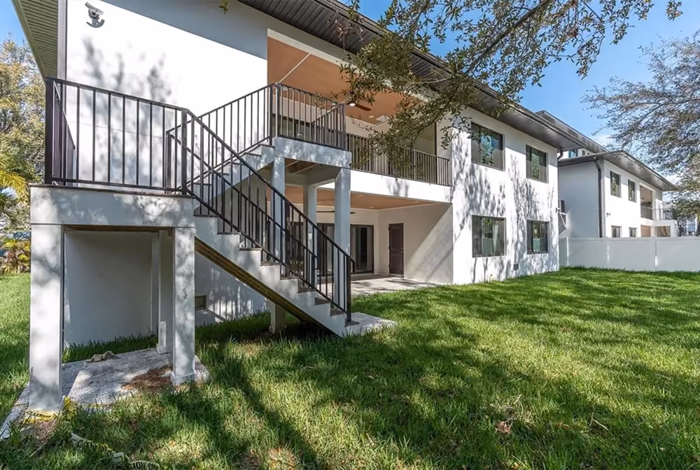 Modern white two-story house with exterior staircase leading to upper balcony and green lawn.