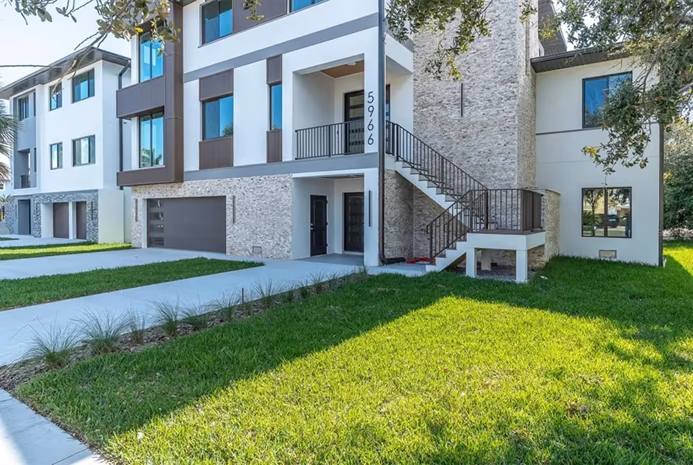 Modern two-story townhouse with garage, exterior stairs, stone accents, and green lawn in front.
