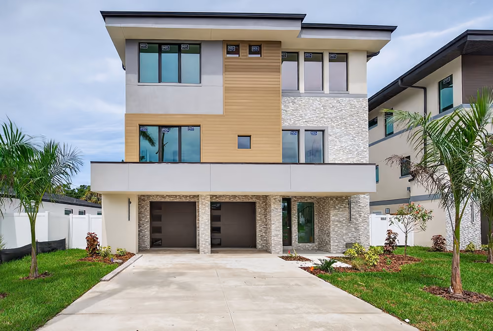 Modern three-story house with stone and wood panel facade, two garage doors, and palm trees in yard.